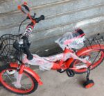 Red kids' bicycle with training wheels and baskets front and rear, wrapped in plastic, leaning against a metal shutter.
