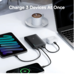 Multi-port charging hub on a white desk charging three devices: a smartphone on the left, a phone at the bottom, and a wireless earbuds case; a laptop is nearby.