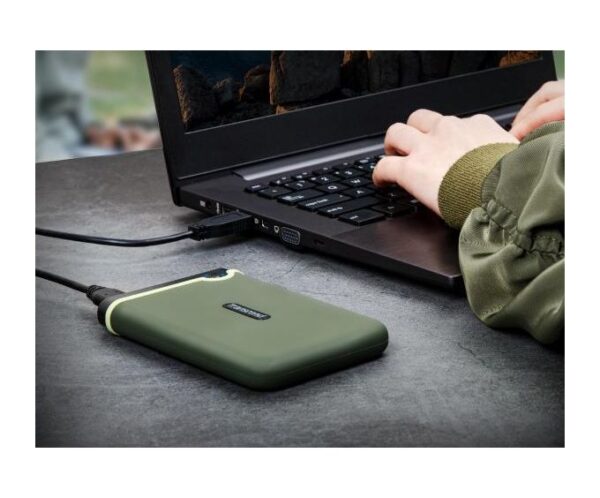 Green rugged external hard drive connected to a laptop via USB as someone types on the keyboard on a gray table.
