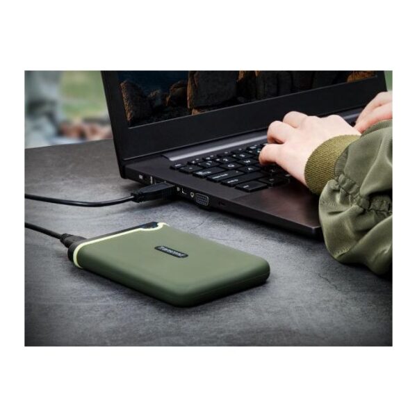 Green rugged external hard drive connected to a laptop via USB as someone types on the keyboard on a gray table.