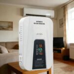 White automatic voltage regulator placed on a wooden table in a living room, with a wall-mounted AC unit above and a sofa nearby.