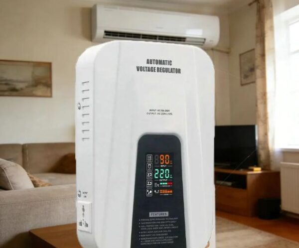 White automatic voltage regulator placed on a wooden table in a living room, with a wall-mounted AC unit above and a sofa nearby.