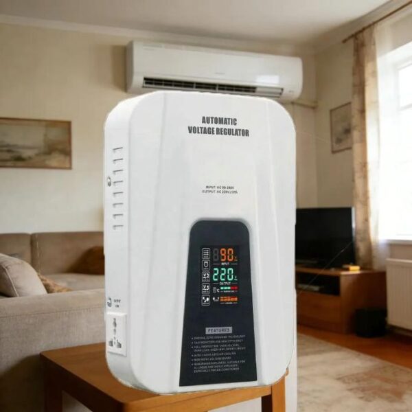 White automatic voltage regulator placed on a wooden table in a living room, with a wall-mounted AC unit above and a sofa nearby.