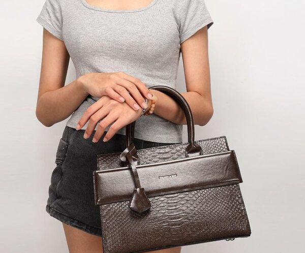Person wearing a gray top and black shorts holding a dark brown textured handbag with top handles and a small front flap pocket nearby.