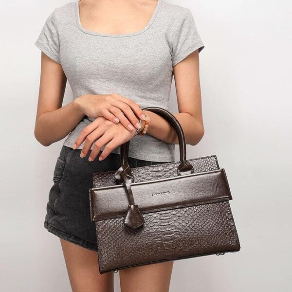 Person wearing a gray top and black shorts holding a dark brown textured handbag with top handles and a small front flap pocket nearby.