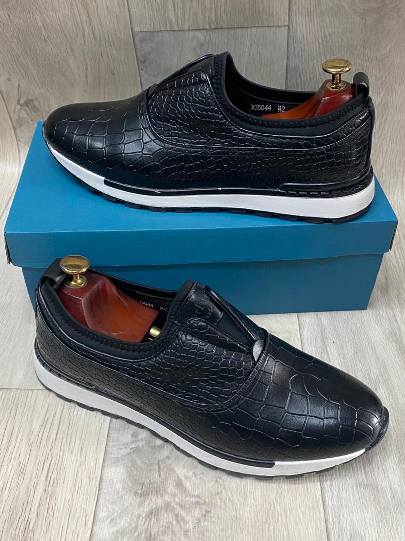 Black crocodile-embossed slip-on sneakers with white cushioned sole, displayed on a blue shoe box.