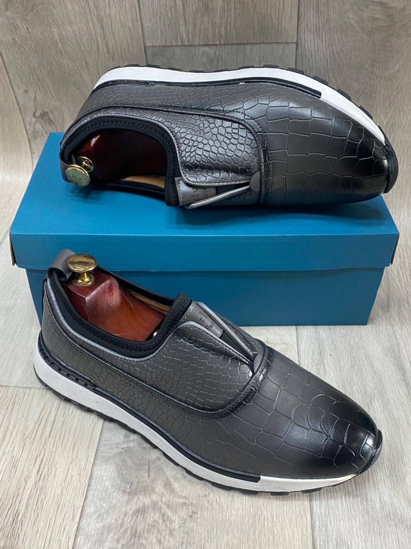Grey crocodile-textured slip-on sneakers with white soles resting on a blue box.