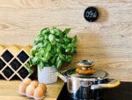Eggs in a carton on a wooden kitchen counter next to a leafy basil plant and a shiny metal pot with a wooden lid.