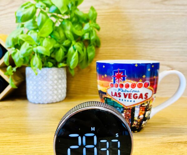Plant in a gray patterned pot beside a colorful Las Vegas souvenir mug on a wooden desk, with a round digital clock in the foreground.