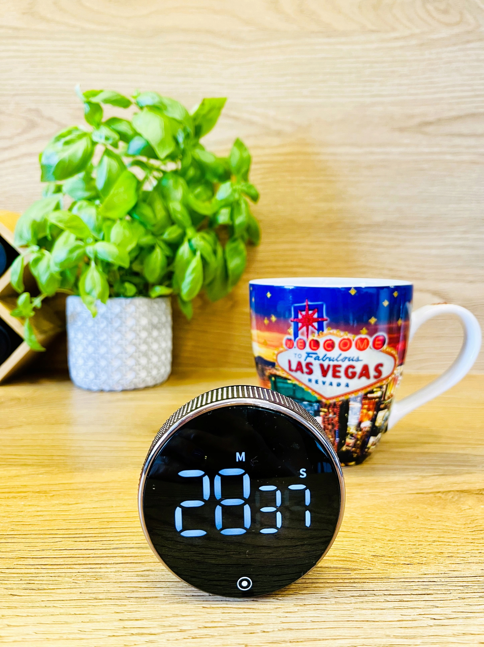 Plant in a gray patterned pot beside a colorful Las Vegas souvenir mug on a wooden desk, with a round digital clock in the foreground.