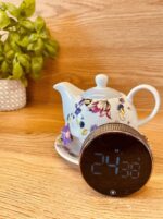 Floral porcelain teapot on a saucer with a round digital alarm clock in front, a green potted plant on the left on a wooden surface.