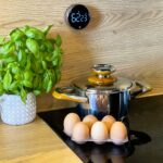 Fresh basil plant in a patterned gray pot sits on a wooden counter beside a stainless steel pressure cooker with wooden handles and a carton of brown eggs in the foreground.