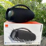 Black Bluetooth speaker with a handle rests on top of its box outdoors, with greenery in the background and a splash image on the packaging.