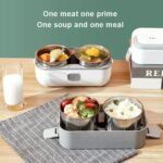 Electric lunch box with multiple metal compartments opened to reveal rice, vegetables, and meat on a wooden table; a glass of milk nearby.