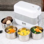 White electric lunch box on a marble counter surrounded by five stainless steel food containers with mixed meals like corn, broccoli, and vegetables.