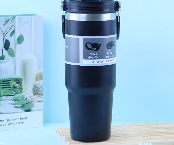 Black insulated travel mug with a flip-top lid and gray instruction band, standing on a wooden surface against a light blue background.