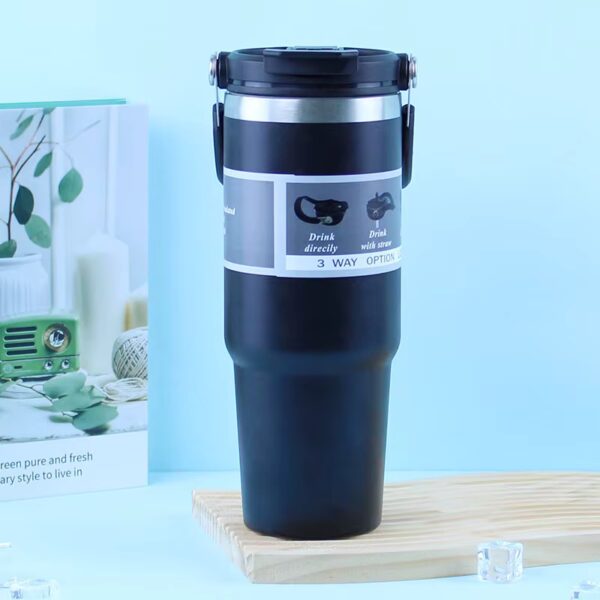 Black insulated travel mug with a flip-top lid and gray instruction band, standing on a wooden surface against a light blue background.